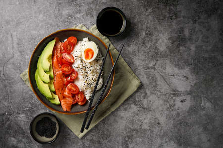 Poke bowl - green bowl with white rice, smoked salmon, cherry tomatoes and avocado, sesame seeds, soy sauce and chopsticks on a gray concrete surface. Top view, negative spaceの写真素材