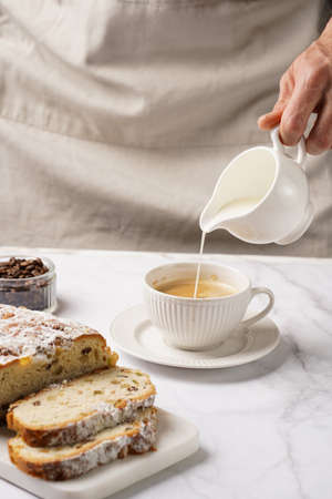 Man pouring milk into a white porcelain cup with coffee, christmas pastry stollen on a marble table