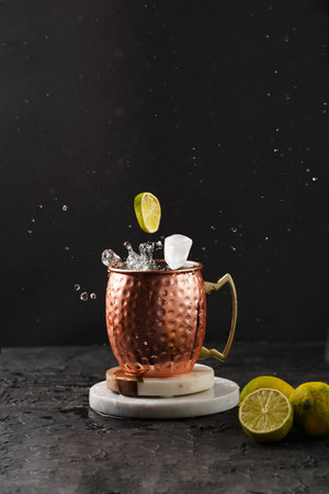 Traditional american alcoholic beverage moscow mule in copper mug with flying lemon and ice cube on white marble boards, black backgroundの写真素材