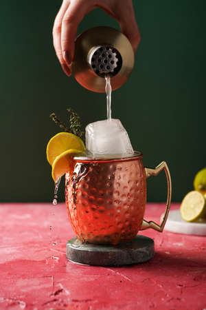 Pouring traditional american alcoholic beverage moscow mule in copper mug with lemon, thyme and huge ice cube from cocktail shaker on magenta and green backgroundの写真素材