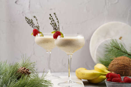Cocktail glasses with sweet banana milk shake beverage, raspberries and coconut on white marble...