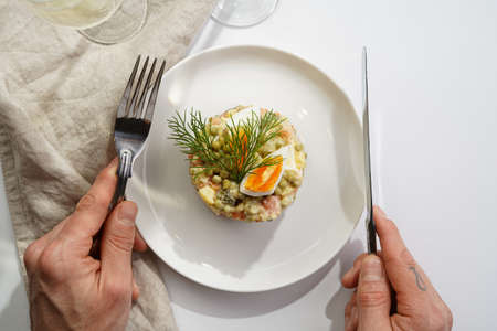 Male hand holding silver fork and knife over a traditional east european dish for New Year's Eve - salad "Olivier" with potatoes, carrots, onion, peas, sausage, cucumbers, eggs and mayonnaise on a white plateの写真素材