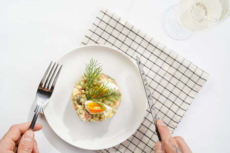 Male hand holding silver fork and knife over a traditional east european dish for New Year's Eve - salad "Olivier" with potatoes, carrots, onion, peas, sausage, cucumbersの写真素材
