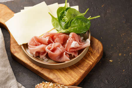 Savory breakfast ingredients: brown lye bread sticks garnished with oats, pork slices prosciutto, hard cheese gouda and fresh greens on wooden boardsの写真素材