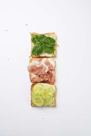 White toasts with cream cheese, ham, cucumbers and spinach on a white surface position in lineの写真素材