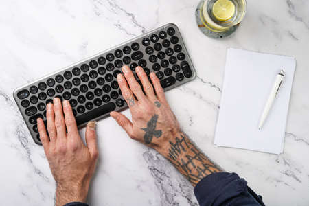 Tattooed male hands working on a wireless keyboard, glass with lemon water, notebook with pen on...