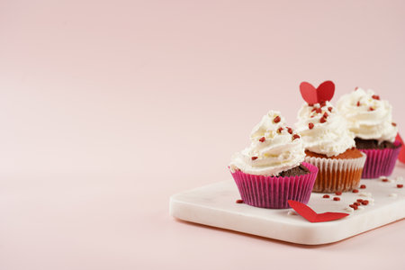 Three homemade white and chocolate muffins with white creamy frosting and heart-shared red and white sugar decoration on marble board on pink surface, copy spaceの写真素材