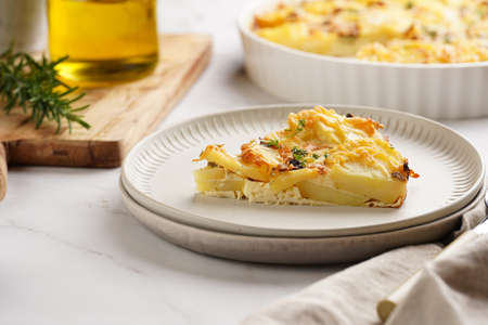Slice of potato casserole with red onions, fresh thyme and rosemary on green checkered kitchen towel, wooden board with flavoring ingredients: pepper, salt, olive oilの写真素材