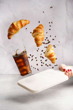 A glass with black iced coffee, coffee beans and freshly baked croissants flying in the air on white...