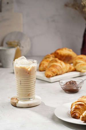 a glass with milk coffee and ice cubes, and freshly baked croissants on marble plate, brown hazelnut...