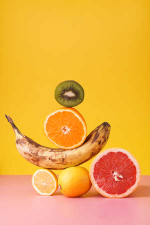 Fruit in balance: dark banana, lemon, grapefruit, kiwi, orange on pink surface on yellow backgroundの写真素材