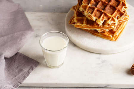 Sweet breakfast - fresh homemade belgian waffles with honey and powdered sugar stacked on white round marble board and small glass with milkの写真素材