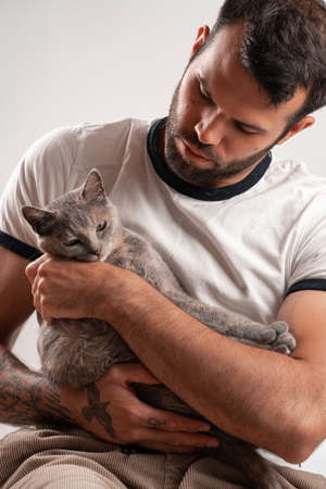 Young tanned Caucasian male with tattoos holding affectionately an adult european short hair cat blue tortie in his armsの写真素材