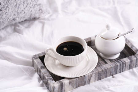 a white porcelain cup with black coffee americano and saucer with sugar in gray marble tray on white bed sheets among fluffy pillows - breakfast in bedの写真素材