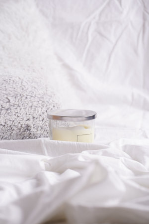 Blank off-white pillar candle in glass jar with label and silver colored lid on white bed sheets and fluffy gray pillowsの写真素材