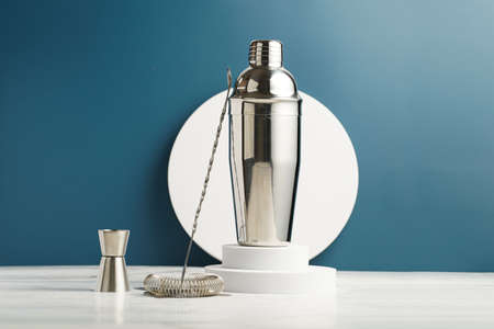 Bartender equipment - stainless steel silver colored cocktail shaker, hawthorne strainer, pourers, bar spoon, muddler , in a bamboos stand on white table on blue backgroundの写真素材