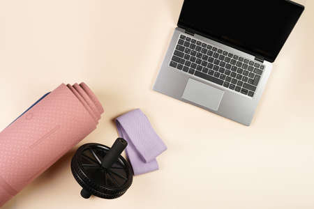 Clothes and equipment for a home workout or gym: pink yoga matt, purple fitness band, black abs wheel, open laptop on beige colored background, top view, copy spaceの写真素材