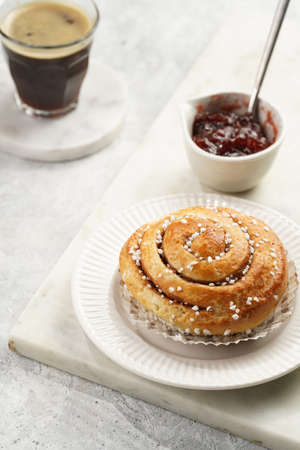 A round cinnamon pastry on a white plate on marble board on light pink grey, cup of coffee, jam, milkの写真素材