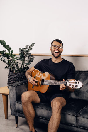 A young white hipster male with glasses, beard and tattoos in black t-shirt sitting on a gray sofa in stylish room against white wall and playing acoustic guitar, looking into the camera cheerfullyの写真素材