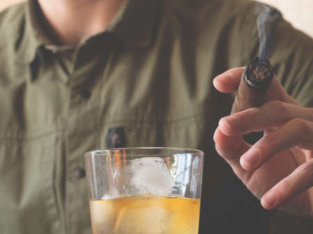 A man in a green shirt holds a Cuban cigar and a glass of whiskey on ice. Close-up of a smoking cigar.の写真素材