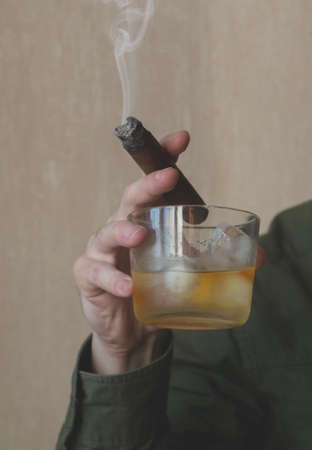 A man in a green shirt holds a Cuban cigar and a glass of whiskey on ice. Close-up of a smoking cigar.の写真素材