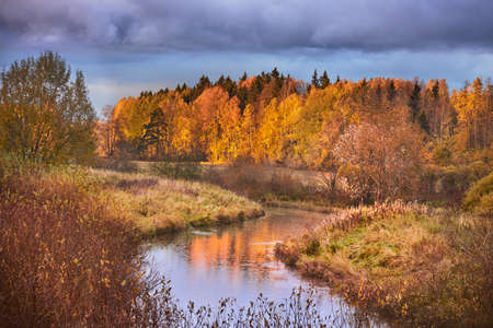 Kerava river flowing between fields and forest in front of colorful autumn background.