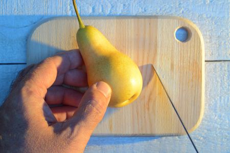 cutting a pear with a hand with a knife on a wooden cutting board on a white wooden table with long blue shadowsの写真素材