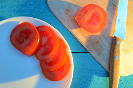 sliced tomato slices on a plate and a wooden cutting board on blue table lit by the sunの写真素材