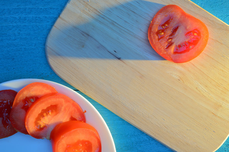 tomato slices are laid out on a wooden cutting board and a white ceramic plate with a blue-turquoise table illuminated by the sun with long cold shadowsの写真素材
