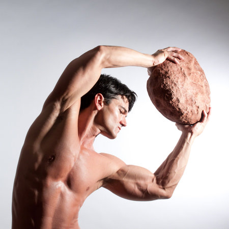 Handsome young man with muscular body and bare torso holding big rock in studioの写真素材