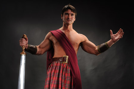 Handsome young man with a muscular body, dressed in a kilt and with a sword on a black backgroundの写真素材