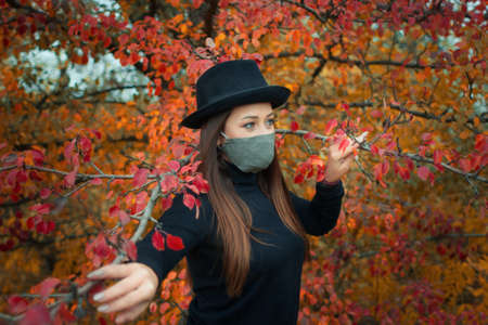 a girl in a protective mask is dressed in a black jacket and hat against the background of an autumn Parkの写真素材