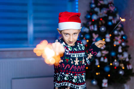 a boy in a Santa Claus hat with sparklers in his hands on the background of a Christmas tree with neon lightinの写真素材