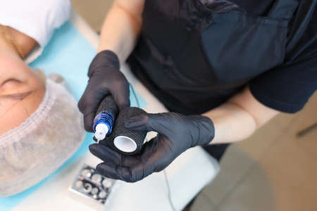 The tattoo artist wraps a special tape for sterility around his device for permanent make-up near the client's faceの写真素材