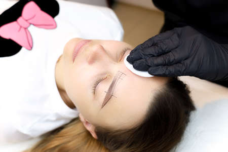a girl who is a master of permanent makeup in a black mask and gloves holds a tattoo machine in her...