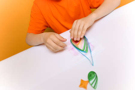 a child in an orange T shirt applies glue to a sheet of white paper to prevent the gluing of thin twisted strips made in the quilling techniqueの写真素材