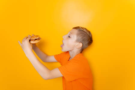 a boy in an orange T shirt holds a hamburger in one hand spreads his arms out to the sides and shows a gesture of bewildermentの写真素材