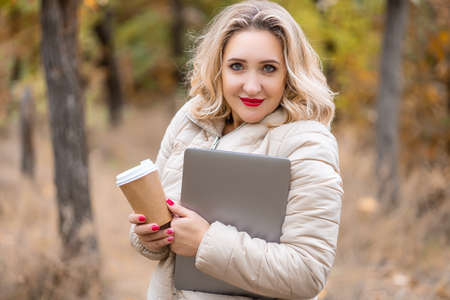 a beautiful girl in an autumn park holds a glass of disposable coffee in her hand and works on a laptopの写真素材