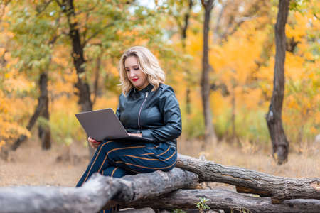 a beautiful girl in an autumn park holds a glass of disposable coffee in her hand and works on a laptopの写真素材