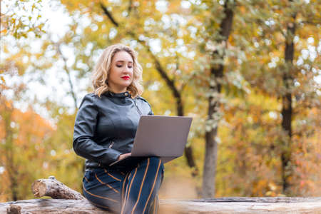 a beautiful girl in an autumn park holds a glass of disposable coffee in her hand and works on a laptopの写真素材