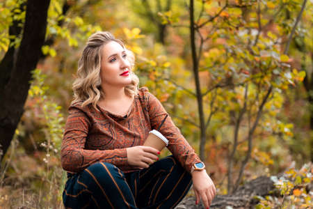 a beautiful blonde is sitting in an autumn park with a glass of coffeeの写真素材