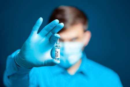 a doctor in a medical mask and gloves dressed in a blue uniform removes the cap from the syringe needleの写真素材