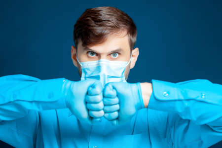 A doctor in a medical mask and gloves, in a blue uniform, poses with thin syringes between his fingers at the cameraの写真素材
