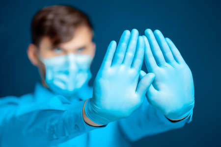 A doctor in a medical mask and gloves, in a blue uniform, poses with thin syringes between his...