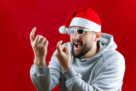 a bearded man in a Santa Claus Christmas hat on a red background imitates an air kiss