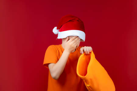 a boy in a Santa's Christmas hat on a red background holds a gift bag in his hand and is very upset