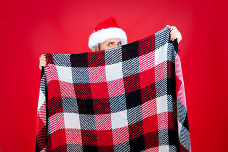 a woman in a red santa hat holds a checkered christmas plaid in front of her and looks out from behind herの写真素材