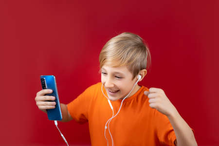 a handsome boy in a yellow T-shirt, who stands on a red background, holds a blue phone in his hand and listens to music on headphonesの写真素材