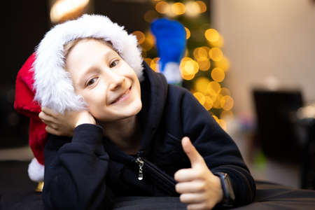 a happy boy in a Christmas hat Santa rolled on his hands and smiles against the background of a Christmas treeの写真素材