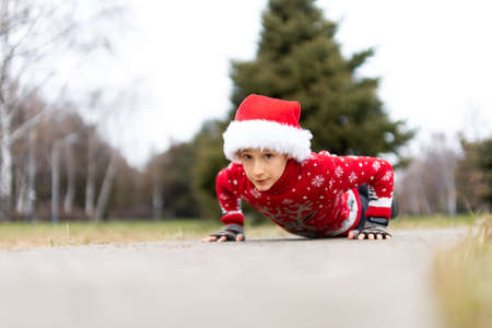 a boy in a warm Christmas sweater with a New Year's deer and a Santa hat performs push-ups in the park on the pathの写真素材